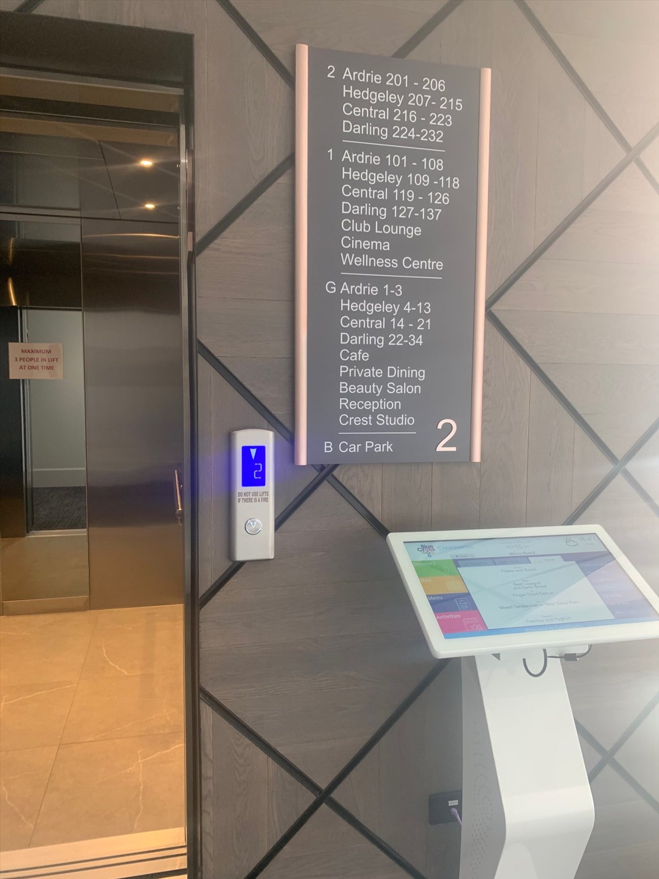A white touch screen kiosk beside a lift with a directory in an aged care home