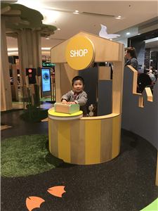A child playing in a shop-themed play kiosk beside a wooden tree at Chadstone