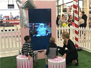 Two children and a helper at a live Santa video booth in a pink gift box